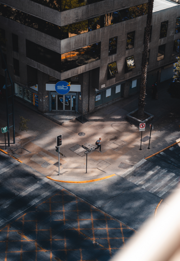 Urban Shadows: Aerial view of a city intersection with a pedestrian crossing the street, shadow patterns on the ground from the surrounding buildings and palm trees.