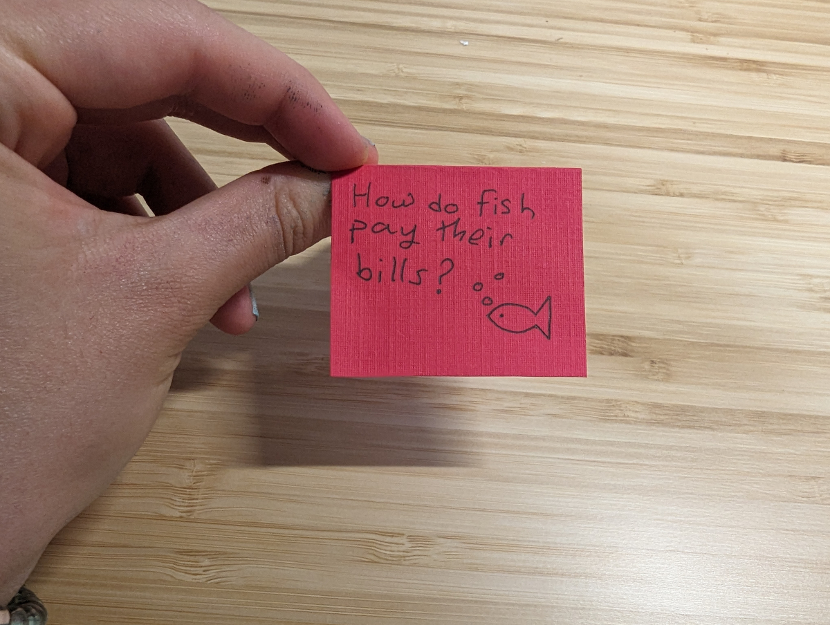 The paper is folded in half with the question 'how do fish pay their bills' written on it.